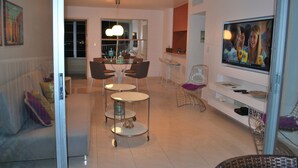 A modern living room with a dining area, a TV, and a glass door leading to a balcony. - Stunning 18th floor remodeled 2/2, beachfront! MONTHLY RENTALS CONSIDERED (Carolina)