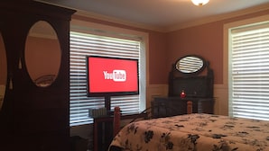 Ein Schlafzimmer mit Bett, Fernseher der YouTube anzeigt, Kommode, Spiegel und Fenster mit Jalousien.
