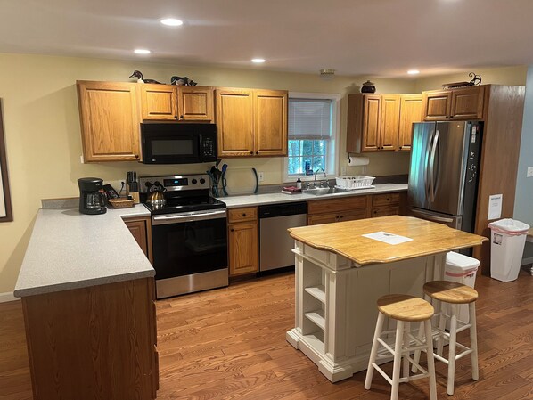 A modern kitchen with wooden cabinets, a stainless steel refrigerator, a built-in oven, and a small island with stools. - Spacious Open Concept House with Beach Access (Moultonborough)