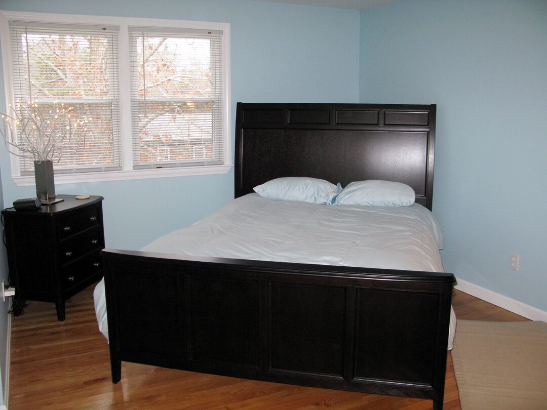 A bedroom with a wooden bed, white bedding, a nightstand, a vase with branches, and a window with blinds.