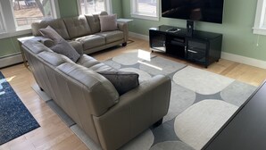 A living room with a beige sectional sofa, a flat-screen TV, and a black entertainment unit. - The Art Nook — Renovated 2BR in the Heart of Downtown Bar Harbor (Bar Harbor)