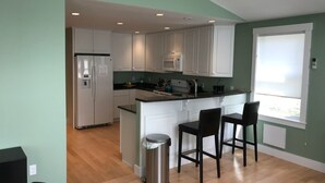 A modern kitchen with white cabinets, a stainless steel refrigerator, a microwave, and a sink. There are two bar stools and a trash can. - The Art Nook — Renovated 2BR in the Heart of Downtown Bar Harbor (Bar Harbor)