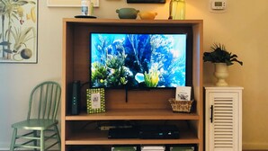 A living room with a television on a wooden stand, a green chair, a white cabinet, and various decorative items on the wall. - Promotions! Stylish Historic District Retreat • Courtyard • Bikes • Walk (Beaufort)