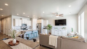 A modern kitchen with white cabinets, a microwave, and a refrigerator. There is a dining area with a wooden table and chairs. A ceiling fan is visible in the living room. - Bright Modern 3BR & Loft - Snow Canyon View w/ Deck (St. George)