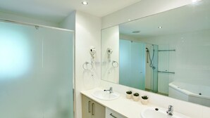 Ein modernes Badezimmer mit zwei Waschbecken, einem großen Spiegel, einer Dusche und Haartrocknern, die an der Wand angebracht sind.