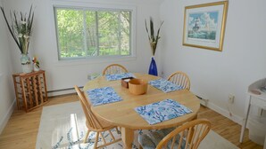 Un comedor con una mesa de madera, individuales azules y sillas. Hay una ventana con persianas, un florero con plantas y un cuadro en la pared.