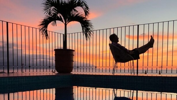 A person relaxing on a balcony with a view of a sunset over water, a palm tree, and a potted plant. - Casa Alexandra. Puerto Vallarta Traditional Comfort. (Puerto Vallarta)