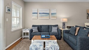 A living room with a grey sofa, striped pillows, a wooden coffee table, and a blue tray. There are three wall-mounted paintings, a lamp, and a small side table. - 12th St / bayside - 4 br multi level townhouse. Nicely furnished and well cared for. (Ocean City)