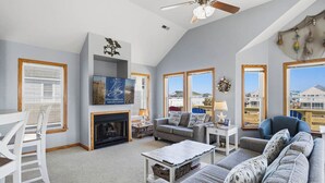 A spacious living room with a fireplace, a television, and a large window offering a view of the neighborhood. - Blue Heron | 955 ft from the Beach | Private Pool, Hot Tub | Nags Head (Nags Head)