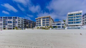 A beachfront property with multiple-story buildings, a sandy foreground, and a clear sky.