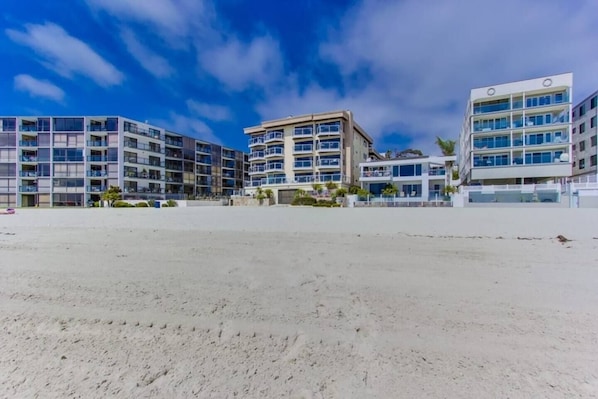 A beachfront property with multiple-story buildings, a sandy foreground, and a clear sky.