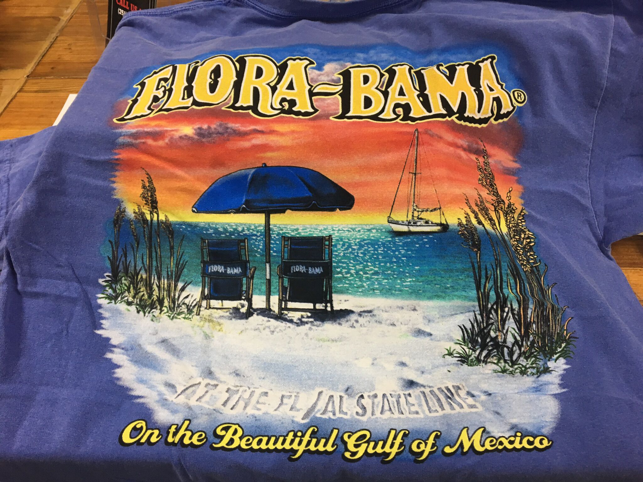 A blue t-shirt with a beach scene, featuring two chairs labeled "FLORA-BAMA" and the text "On the Beautiful Gulf of Mexico."