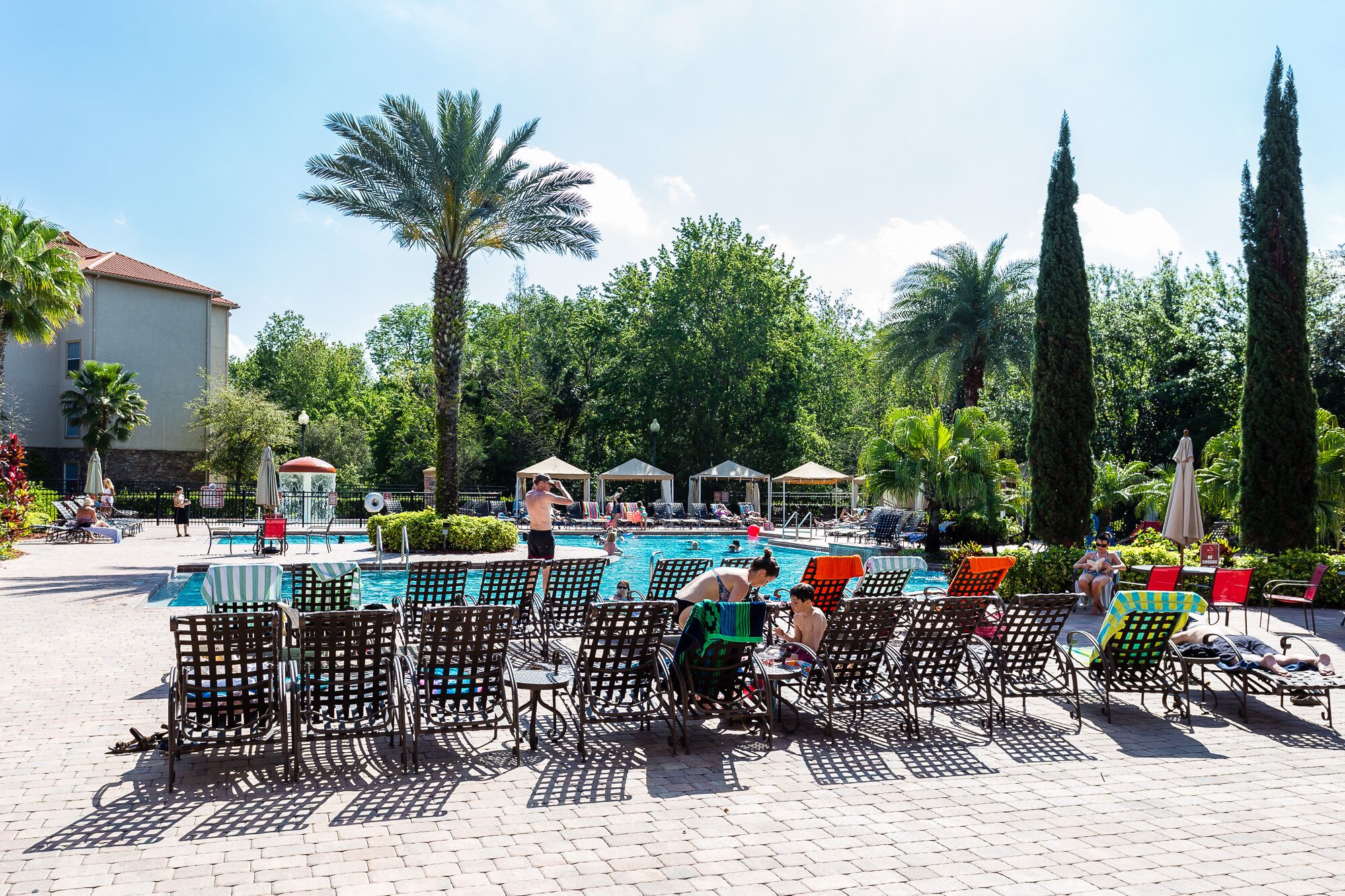 A poolside area with lounge chairs and umbrellas, surrounded by palm trees and a building in the background.