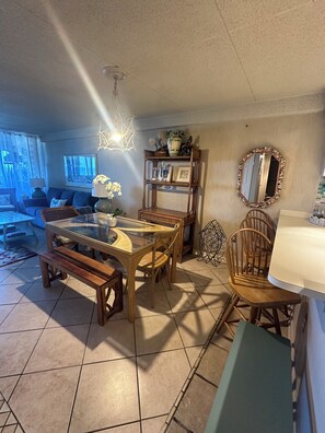 A living room with a glass-top dining table, wooden chairs, a sofa, a TV, and a decorative shelf.