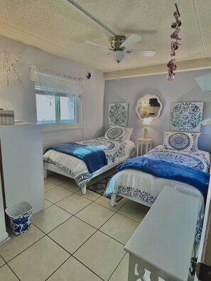 A bedroom with two beds, a ceiling fan, a window with blinds, and decorative wall art.
