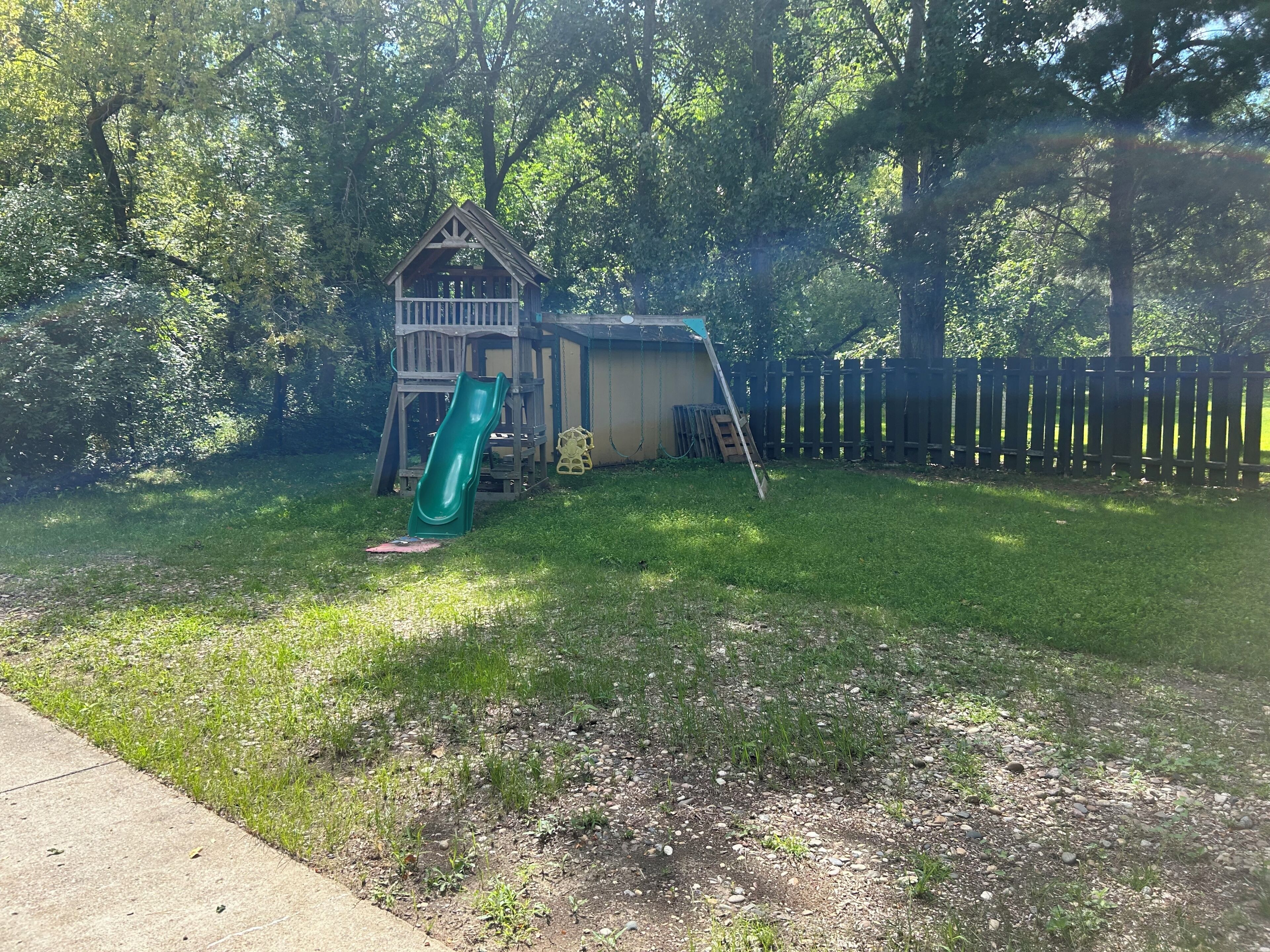 A backyard with a wooden playhouse, a green slide, a ladder, and a fenced area.