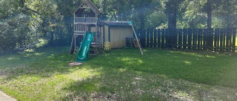 A backyard with a wooden playhouse, a green slide, a ladder, and a fenced area.