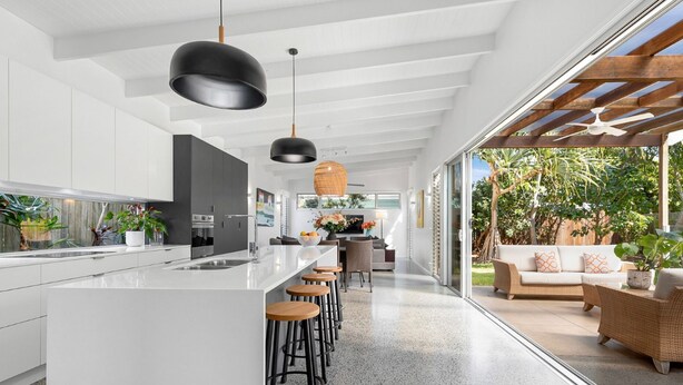 A modern kitchen with white cabinetry, a central island with stools, pendant lighting, and a view of a garden through large sliding doors. - Elanda St - Luxurious Beachside Living (SUNSHINE BEACH)
