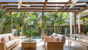 An outdoor patio with wicker furniture, a glass-top coffee table, and a pool in the background. - Elanda St - Luxurious Beachside Living (SUNSHINE BEACH)
