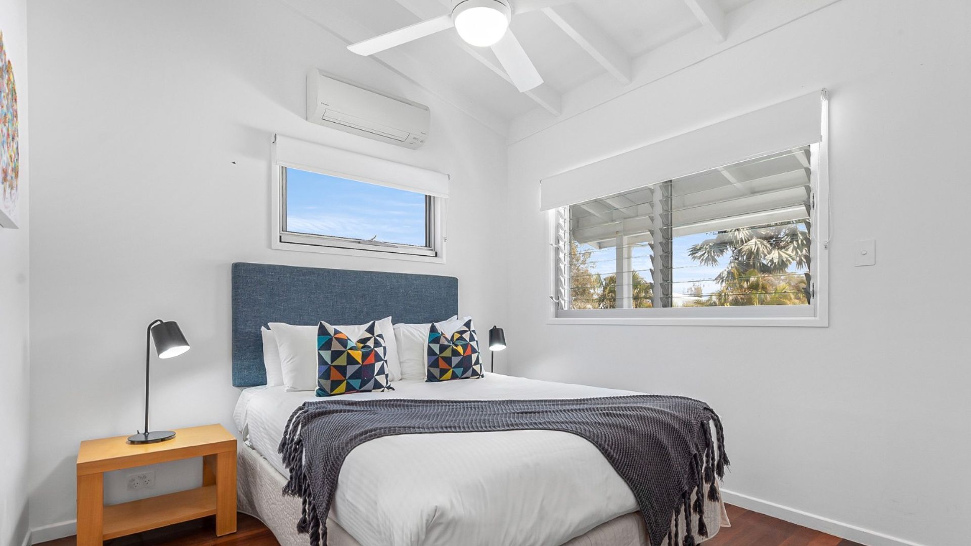 Une chambre à coucher avec un lit, une table de chevet, un ventilateur de plafond et une fenêtre donnant sur des palmiers.
