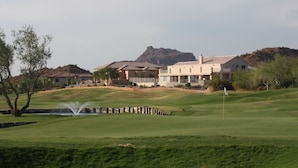 A golf course with a water fountain, a flag, and a building with multiple stories in the background.