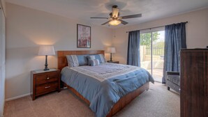 A bedroom with a bed, two bedside tables with lamps, a ceiling fan, a large window with curtains, and a view of the outdoors.