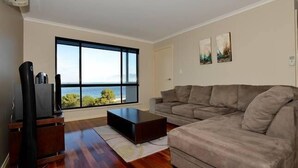 A living room with a large window, a gray sofa, a coffee table, and a TV on a stand. - Wahroonga On The Water (Kingscote)