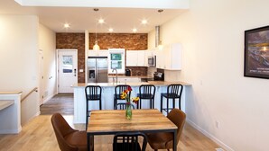 Une cuisine moderne avec des meubles blancs, un mur d’accent en briques et un coin repas comprenant une table et des chaises en bois.