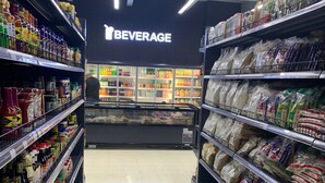 Ein Gang in einem Supermarkt mit Regalen voller Lebensmittel und einem Kühlbereich im Hintergrund, der "BEVERAGE" zeigt.
