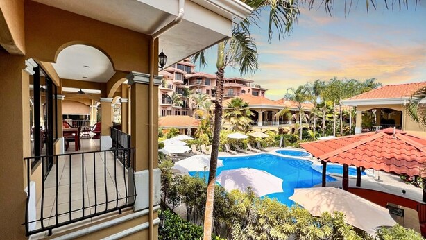 Un balcón de hotel con vistas a la piscina, mobiliario exterior y un jardín tropical.