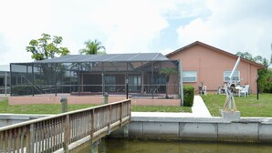 A building with a glass enclosure, a wooden bridge over water, and a grassy area with outdoor furniture. - Home on the water w/ pool and dock (Apollo Beach)