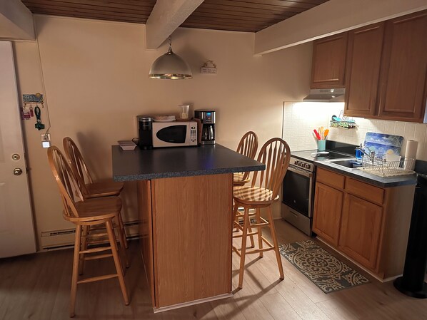 A compact kitchen with wooden cabinets, a central island with chairs, a microwave, a coffee maker, and a stove. - Heated Pool , One Block to Private Beach (Ortley Beach)
