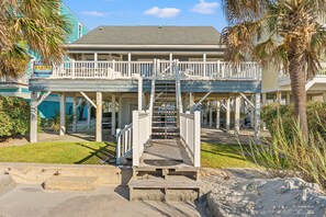 A beachfront property with a wooden deck, a staircase leading to a balcony, and a rental sign.