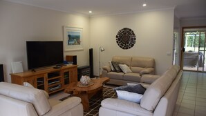 A living room with a flat-screen TV, wooden entertainment unit, beige sofa, and a patterned rug. - Beach House - direct 2-min access to the beach (Depot Beach)