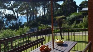 A covered patio with a table and bench, a bowl of fruit, and a view of a grassy area and trees. - Beach House - direct 2-min access to the beach (Depot Beach)
