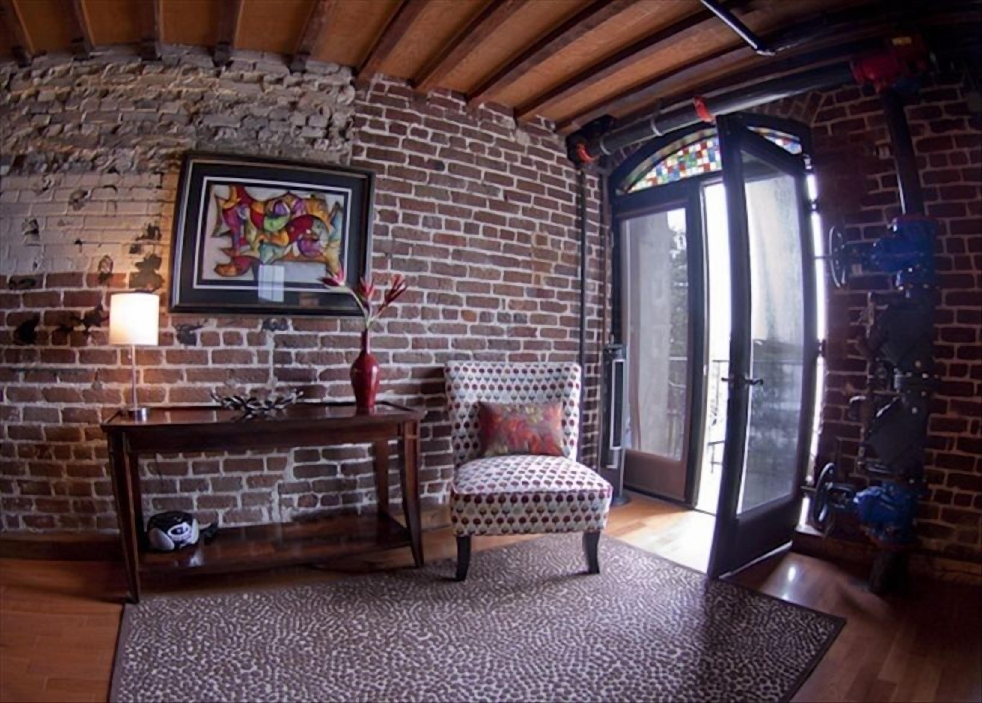 A cozy room with exposed brick walls, a wooden ceiling, a patterned armchair, a small table with a vase, and a door leading outside.