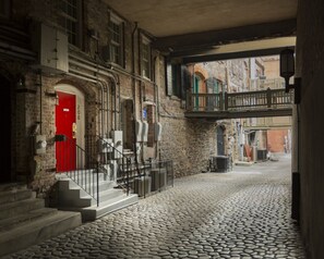 Ein Kopfsteinpflasterhof mit einer roten Tür, einer Treppe und einem Balkon mit Blick auf eine Straße.