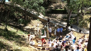 Evento ao ar livre com pessoas sentadas em cadeiras brancas, uma parede de pedra e árvores ao fundo.