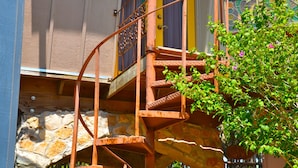 Uma casa com escadaria em espiral que dá para uma varanda, cercada por plantas e um pilar de pedra.