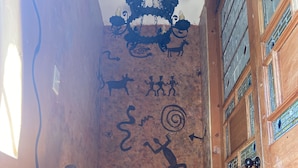 Um corredor estreito com paredes e chão de madeira. Há um candelabro pendurado no teto e uma porta com janela no lado direito.