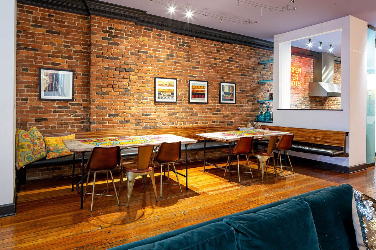 A cozy dining area with wooden tables and chairs, a brick wall with framed pictures, and a neon sign indicating the establishment is open 24 hours.