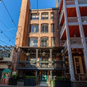En bygning med flere etager og facade af mursten, en café i stueetagen med udendørs siddepladser, og en grøn dør.