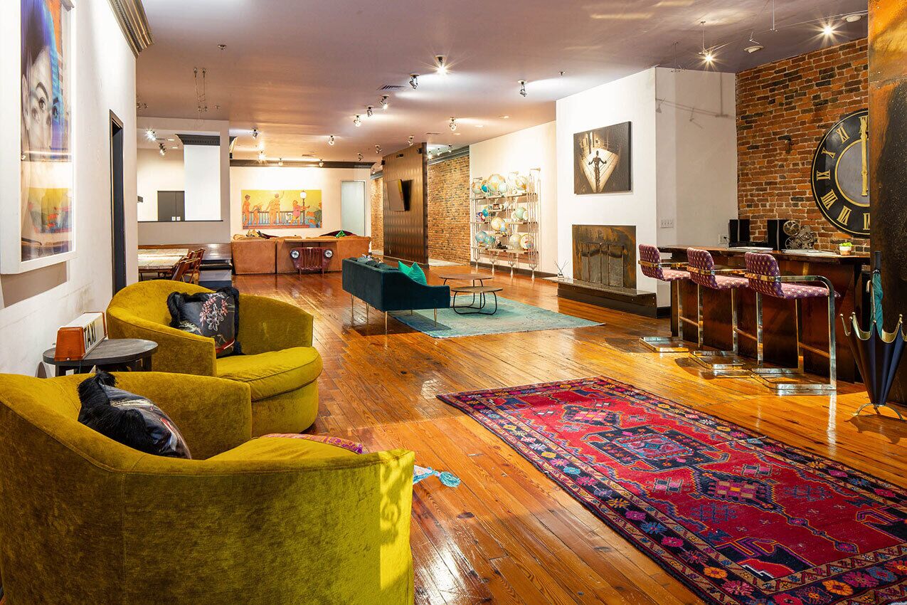 A spacious living area with a wooden floor, yellow armchairs, a colorful rug, a brick wall, and a large clock.
