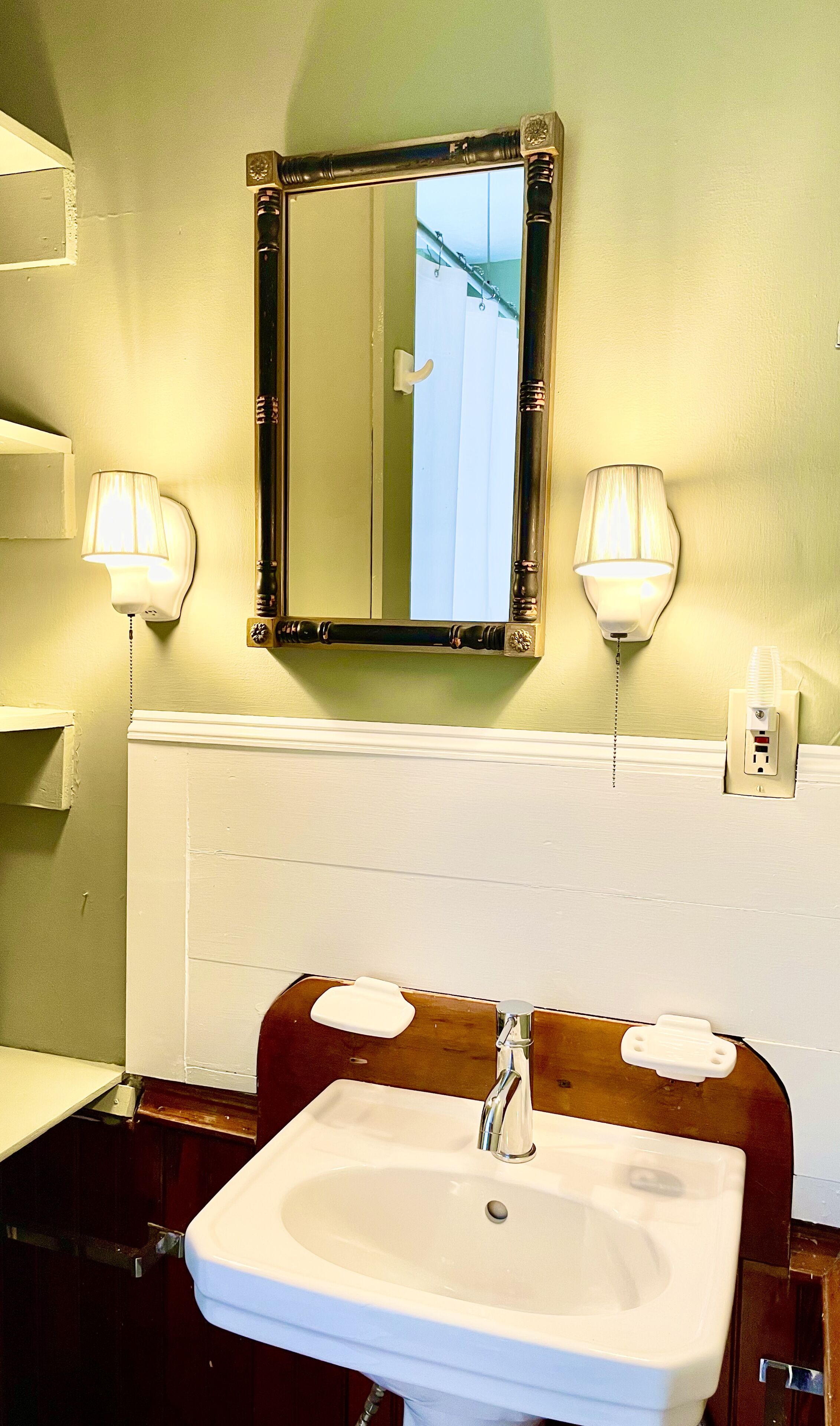 Um banheiro com uma pia branca, duas luminárias na parede e um espelho com moldura decorativa.