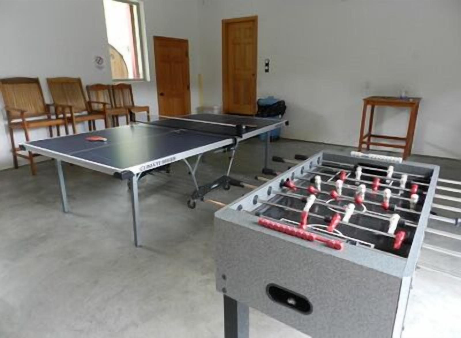 A room with a foosball table, a ping pong table, and several chairs.