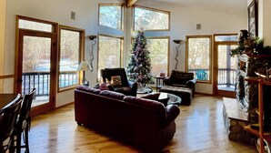 A spacious living room with a wooden ceiling, large windows, a Christmas tree, and a stone fireplace. -  Riverdance Lodge: Best Family and Friends private Icicle River Fun Getaway!! (Leavenworth)