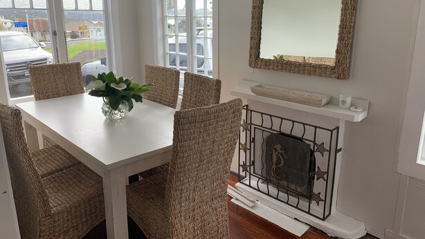 A dining area with a white table, wicker chairs, a vase with flowers, a fireplace with a metal grate, and a mirror with a wicker frame.