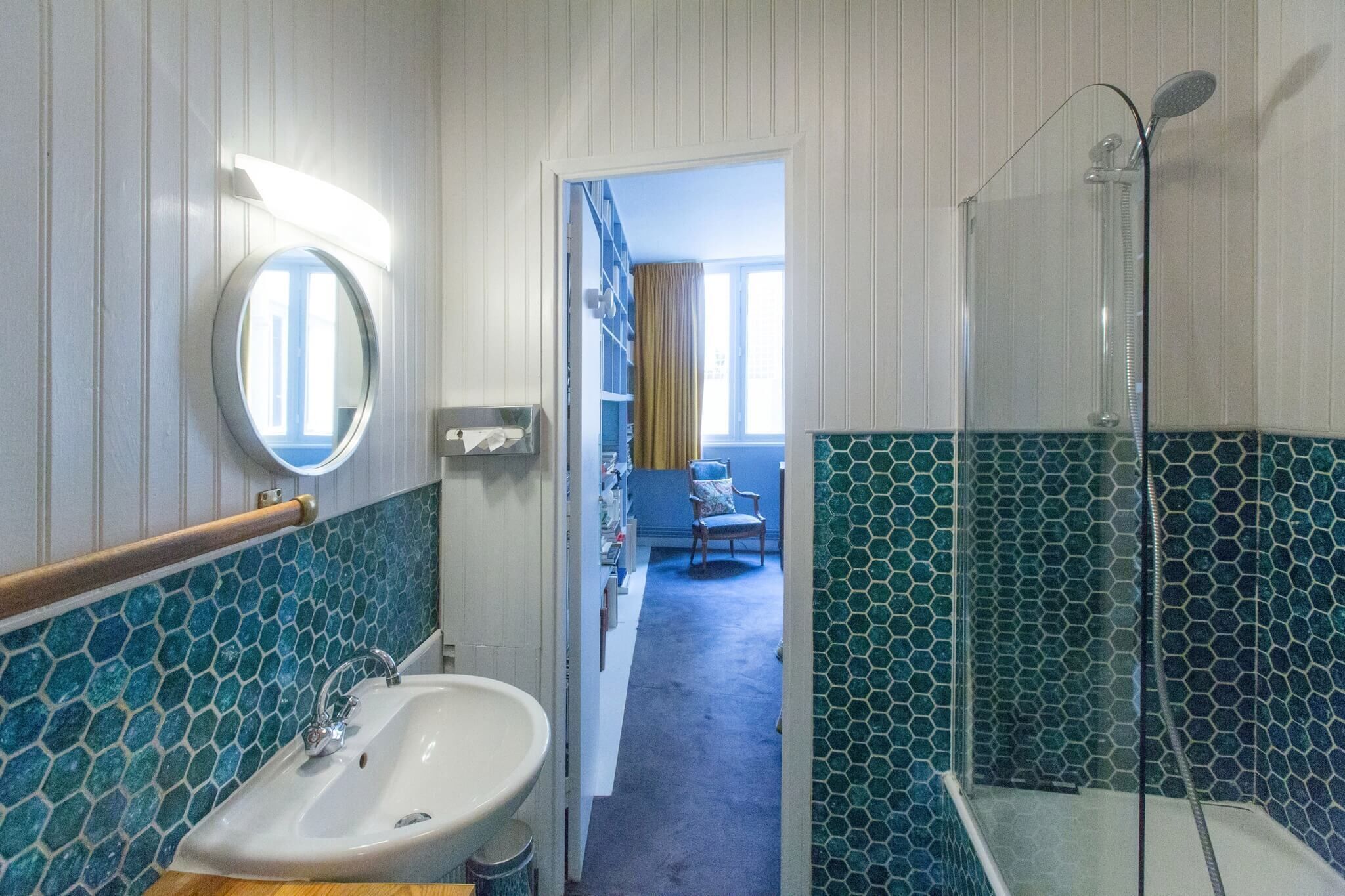Un bagno con lavandino, specchio e doccia. Nell'ambiente adiacente, un tappeto blu e una sedia.
