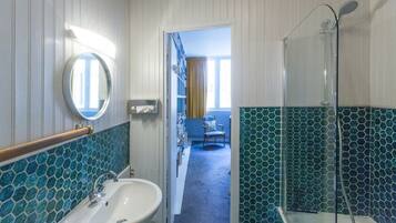 Un bagno con lavandino, specchio e doccia. Nell'ambiente adiacente, un tappeto blu e una sedia.