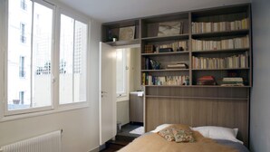 A bedroom with a large window, a bookshelf, and a bed with a blanket and pillow. - Eclectic large living space steps from the the Eifel Tower 2bedrs 2,1/2bathrs (Paris)